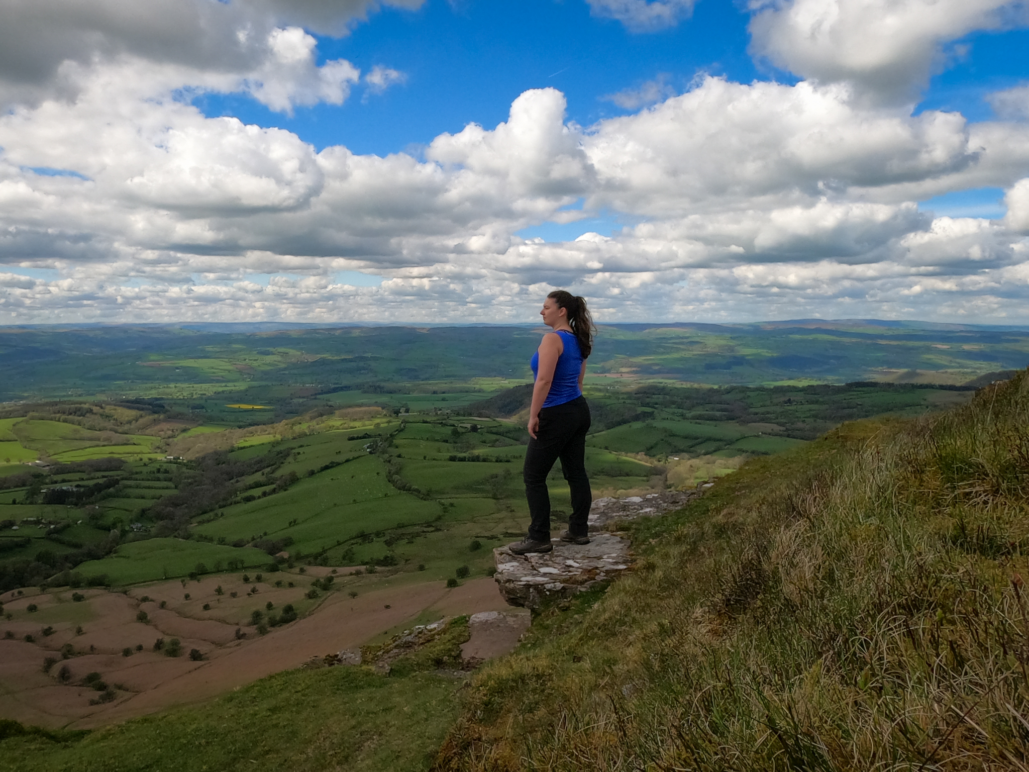 A woman with a long dark pink skirt and turquoise Osprey backpack, with hiking boots tied to it, walking away on a woodland path in the forest surrounded by bluebells.
