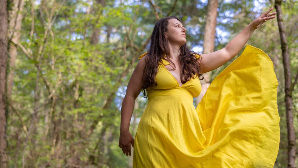 A brunette woman in the woods, with long flowing hair, wearing a long yellow dress. One hand is lifted and the skirt is fluttering in the wind.