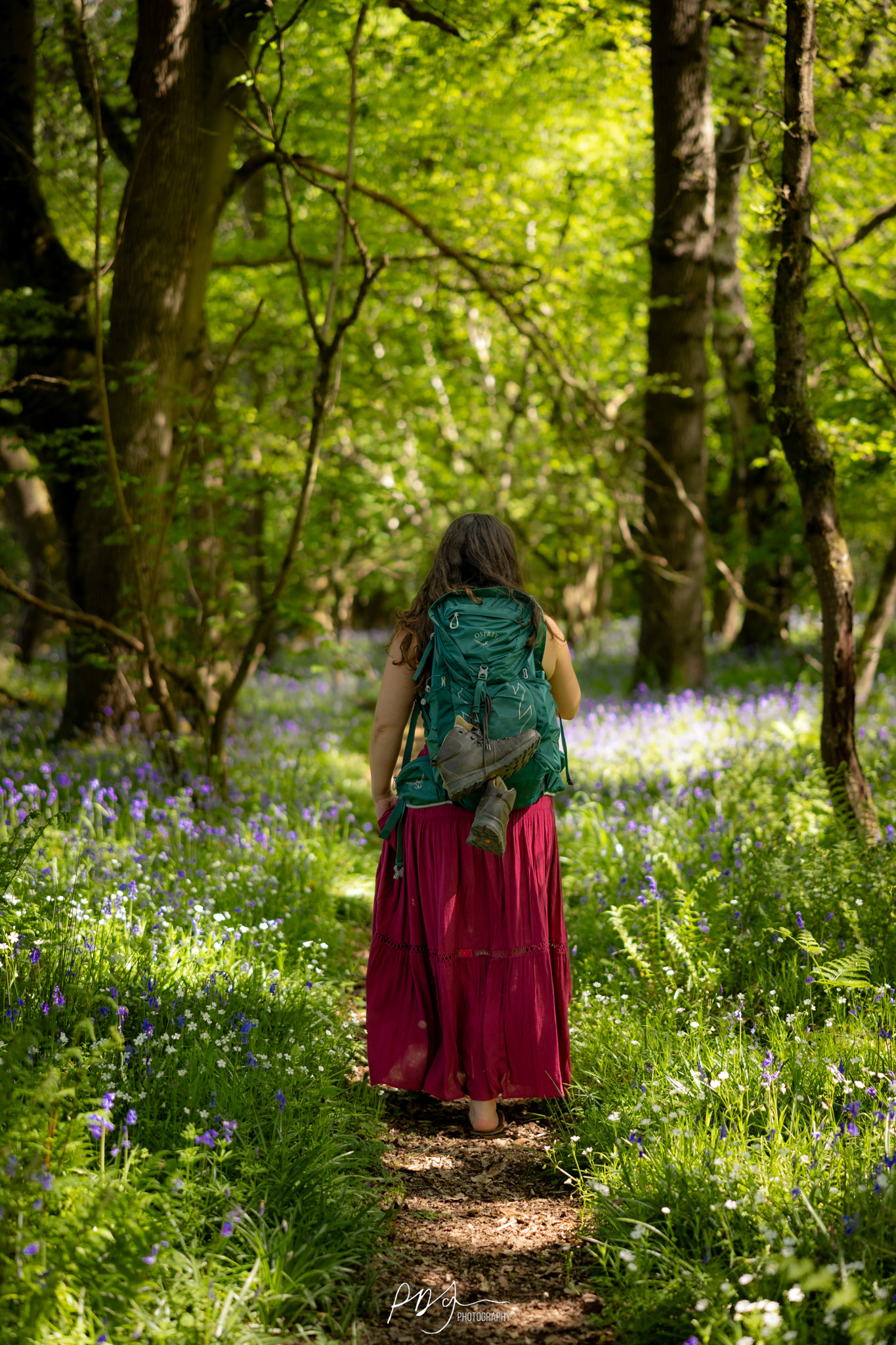 A woman with a long dark pink skirt and turquoise Osprey backpack, with hiking boots tied to it, walking away on a woodland path in the forest surrounded by bluebells.