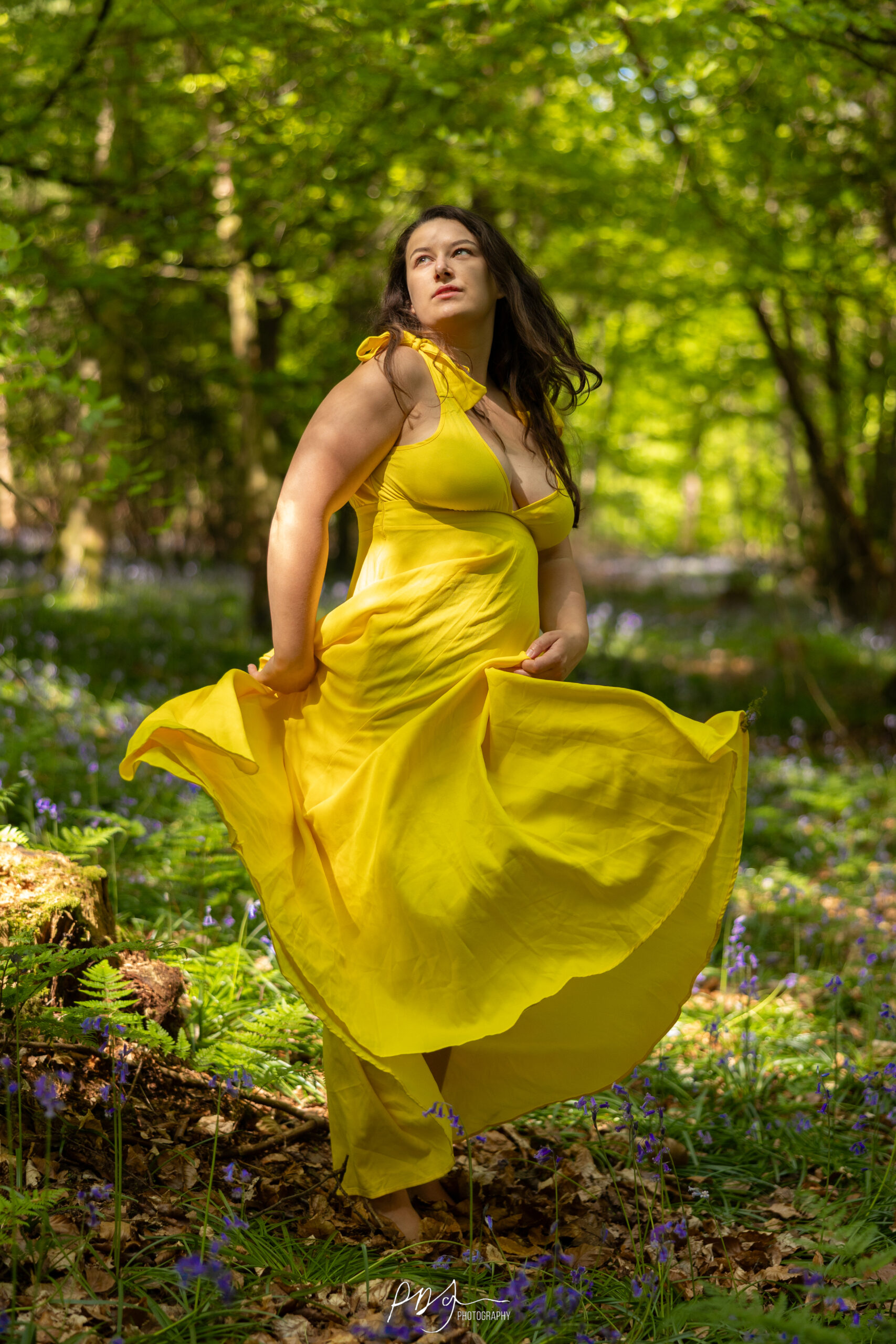A woman in a yellow dress twirling in the spring woods, surrounded by trees and bluebells