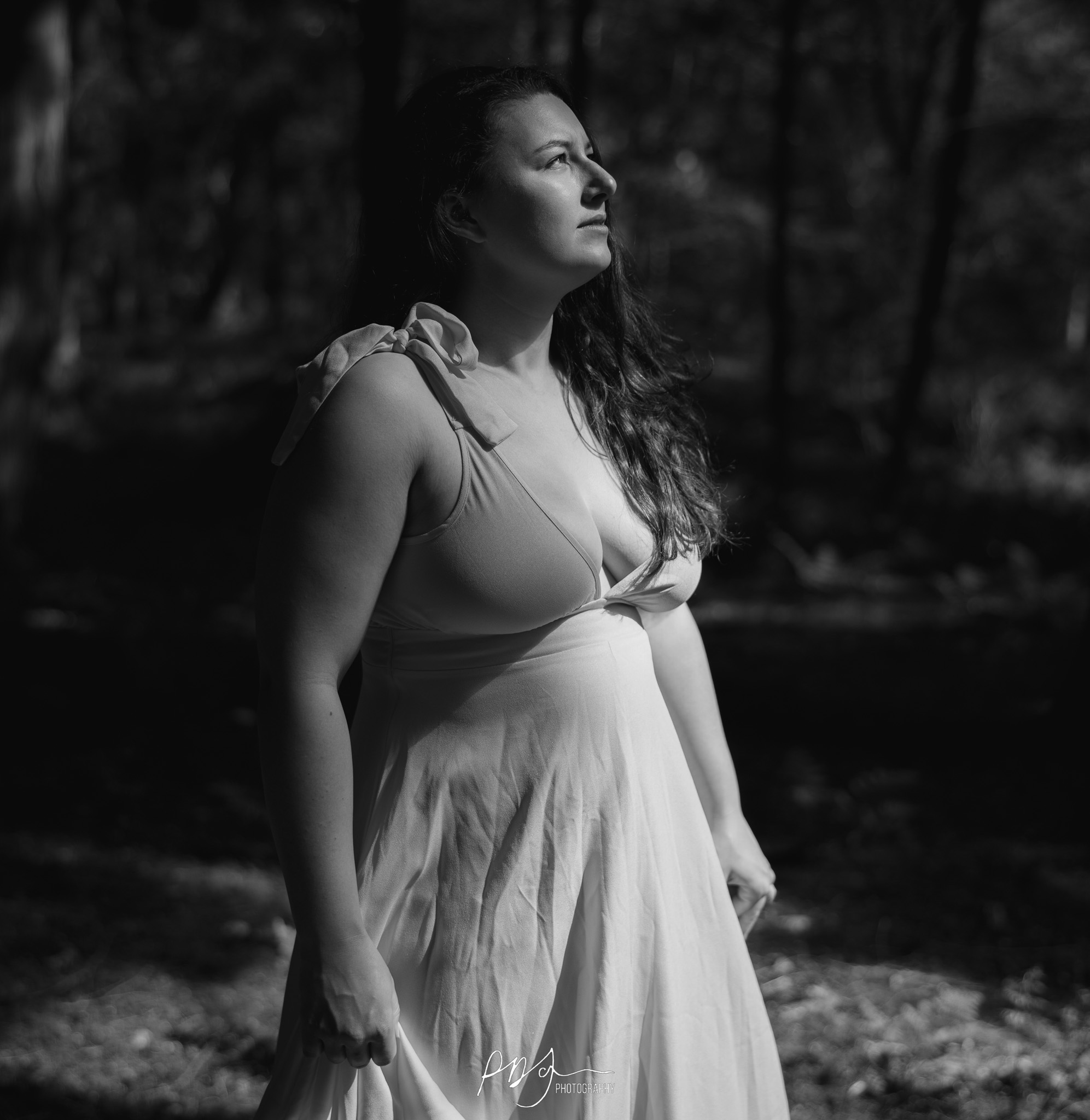 A black and white image of a woman with dark hair waering a long, light-coloured dress looking up into a sunbeam