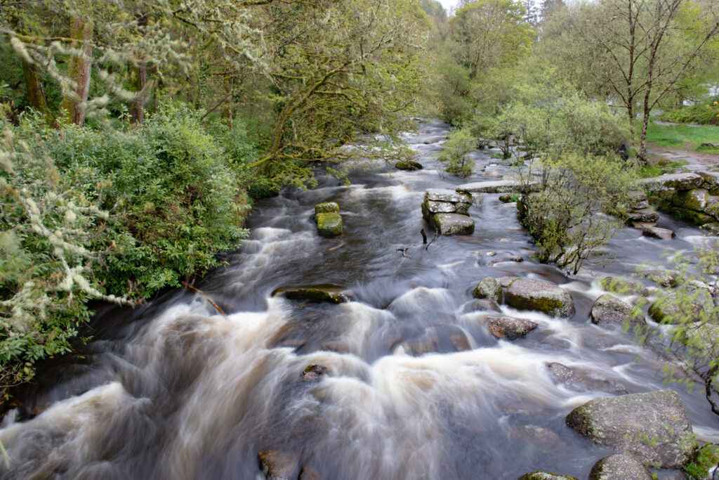 A slow shutterspeed capture of a river full of large boulders, surround by trees covered in lichen, with a partially-standing stone clapper bridge from 1792.